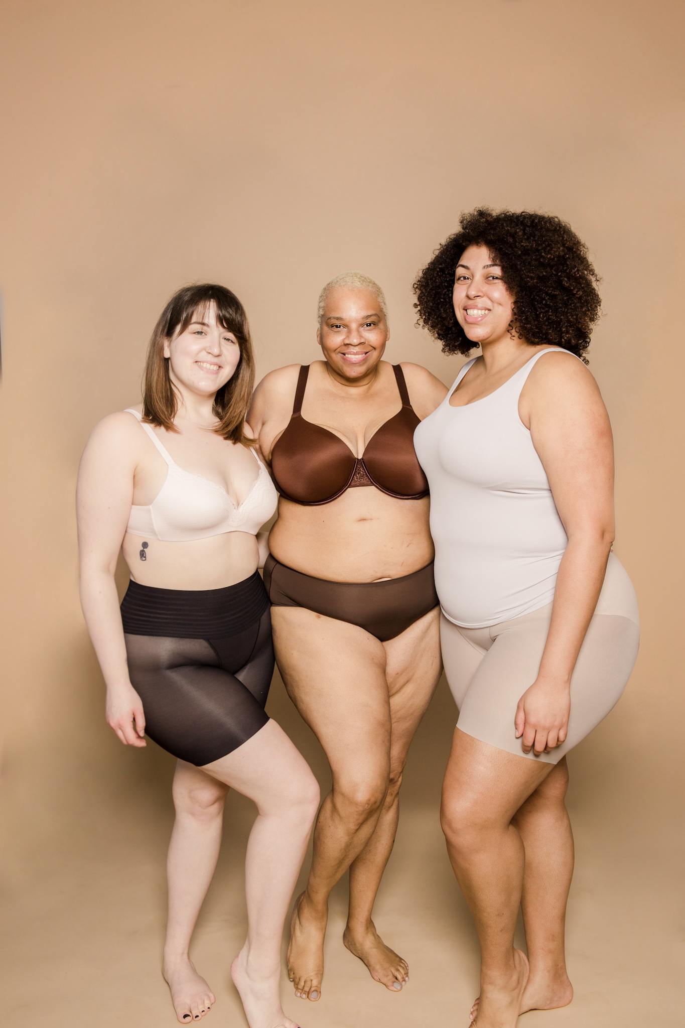 Full body group of cheerful plus size multiethnic female models wearing lingerie standing close and looking at camera on beige background in studio