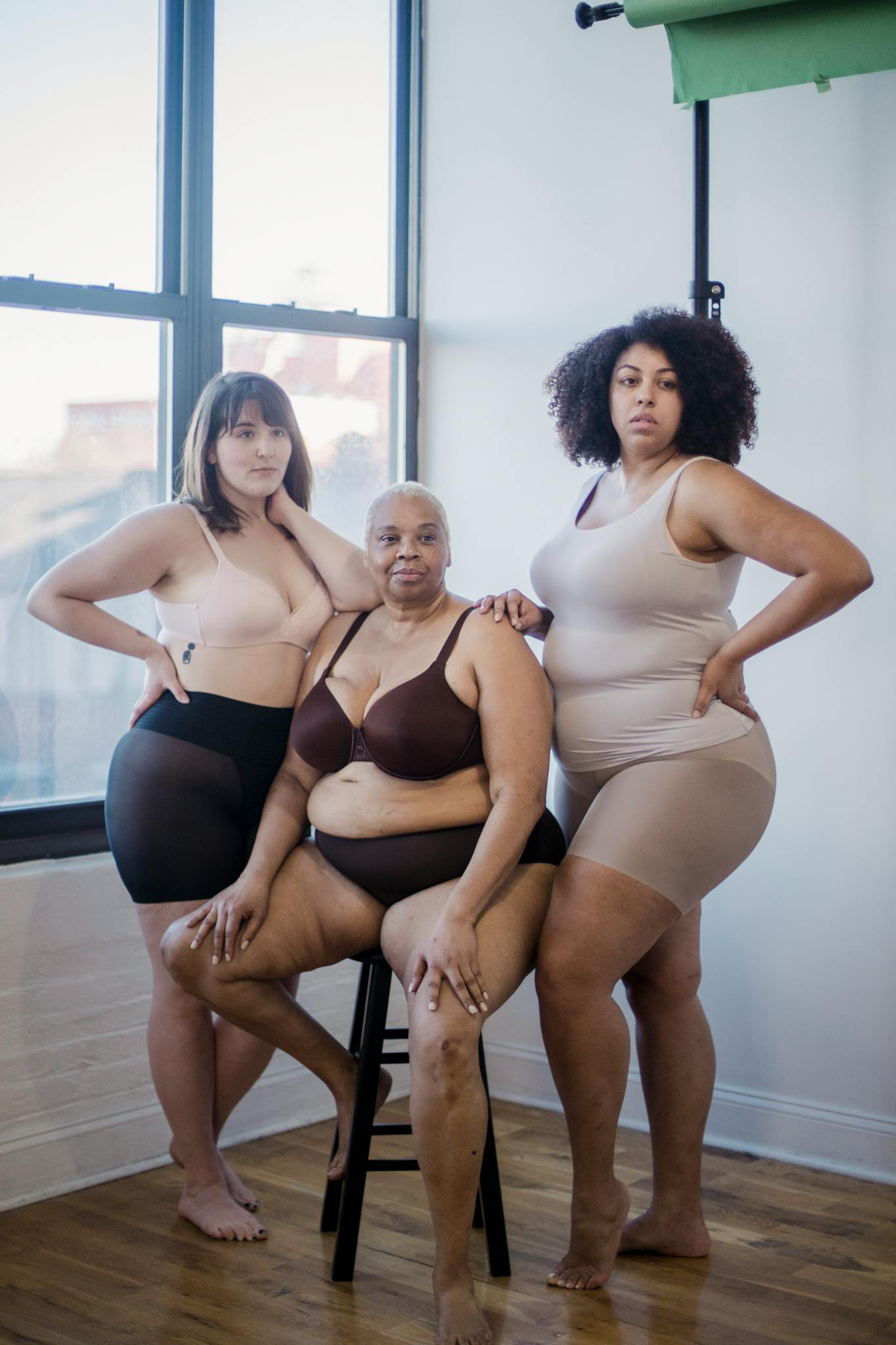 Full body of confident plump multiracial female models wearing lingerie looking at camera while standing near window in light room