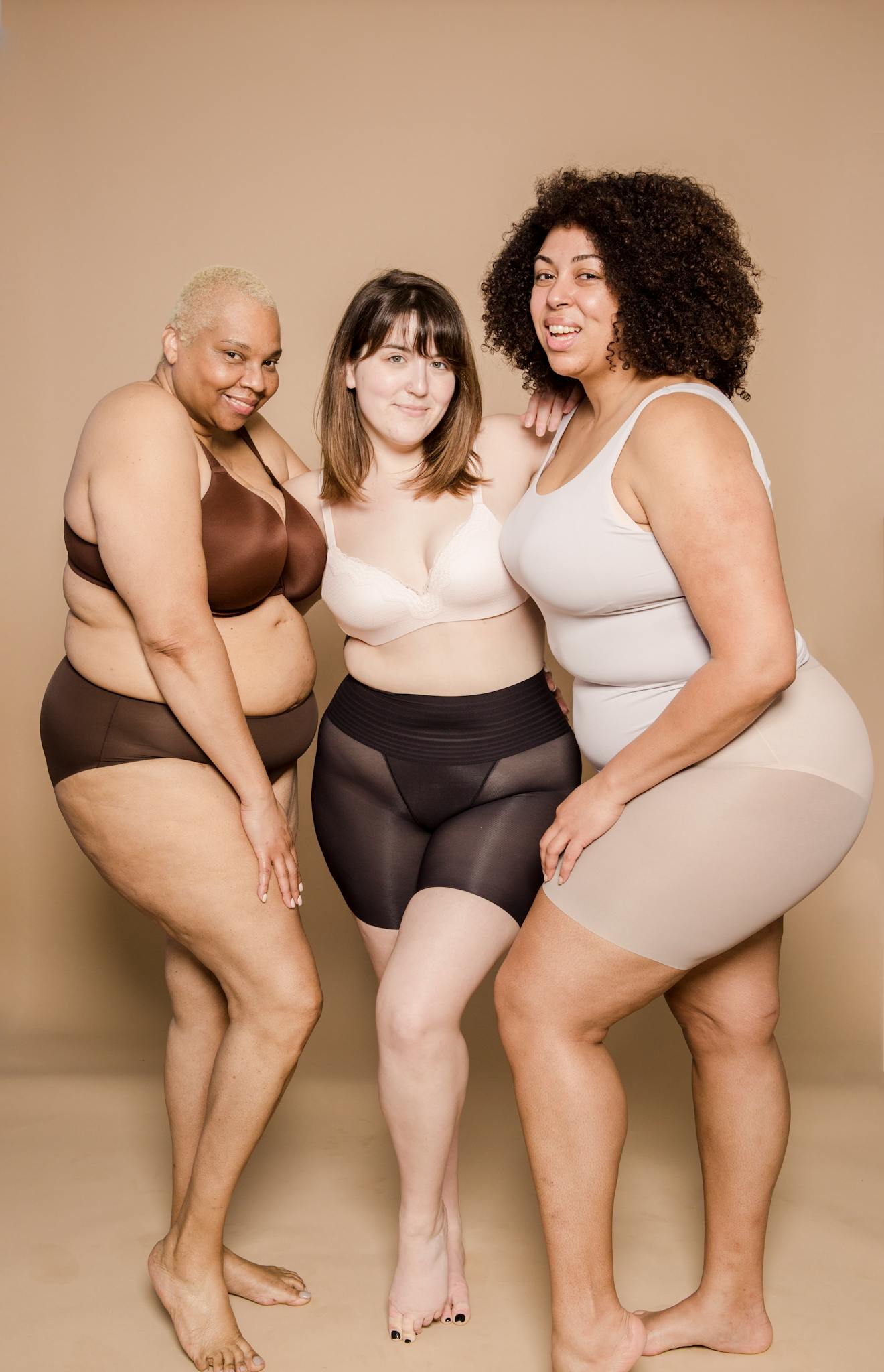 Full body of plump multiethnic barefoot female models wearing underwear looking at camera while standing on beige background in studio