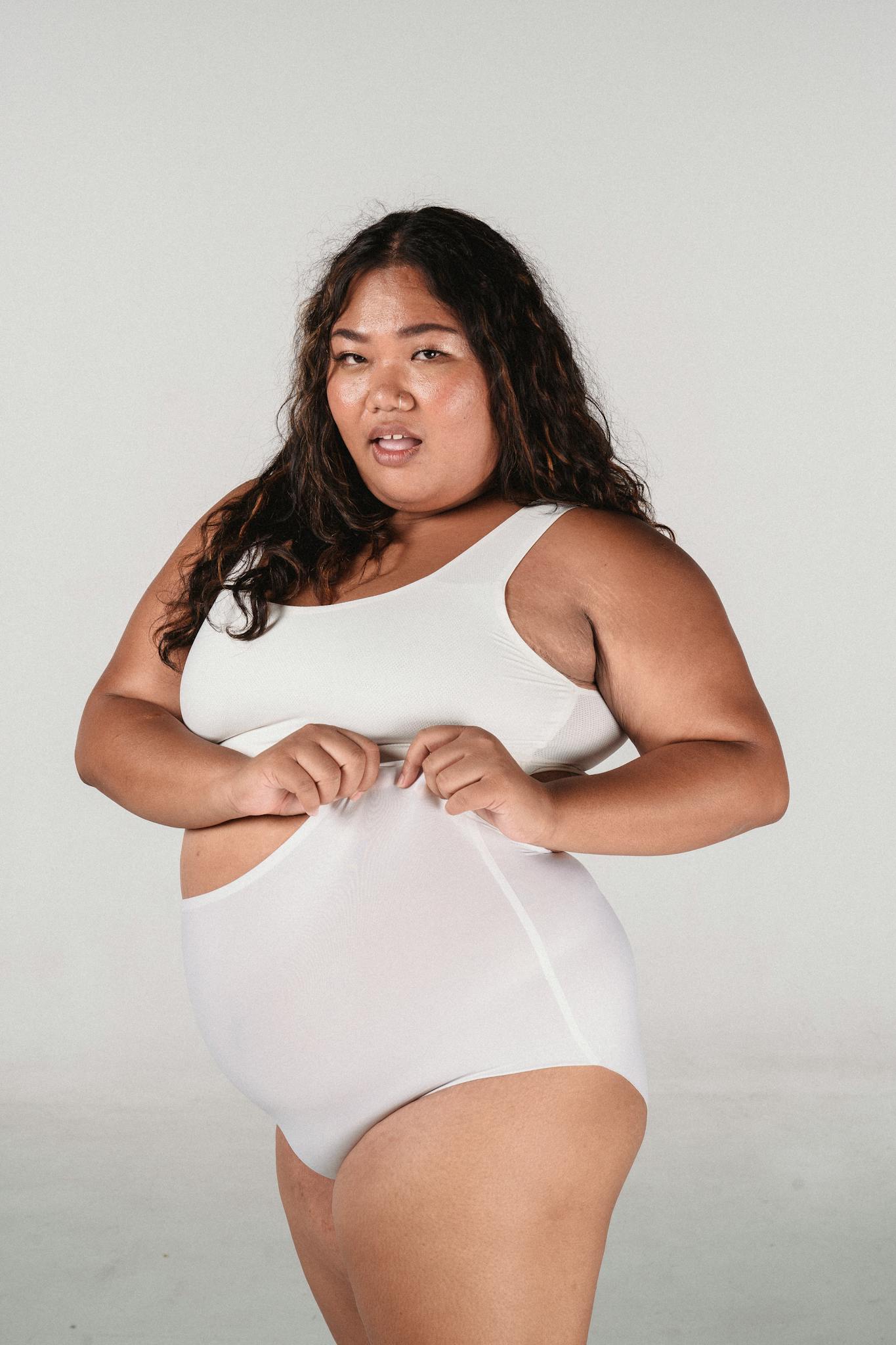 Dreamy plus size Asian female looking at camera wearing modeling panties and top while standing on white background in studop