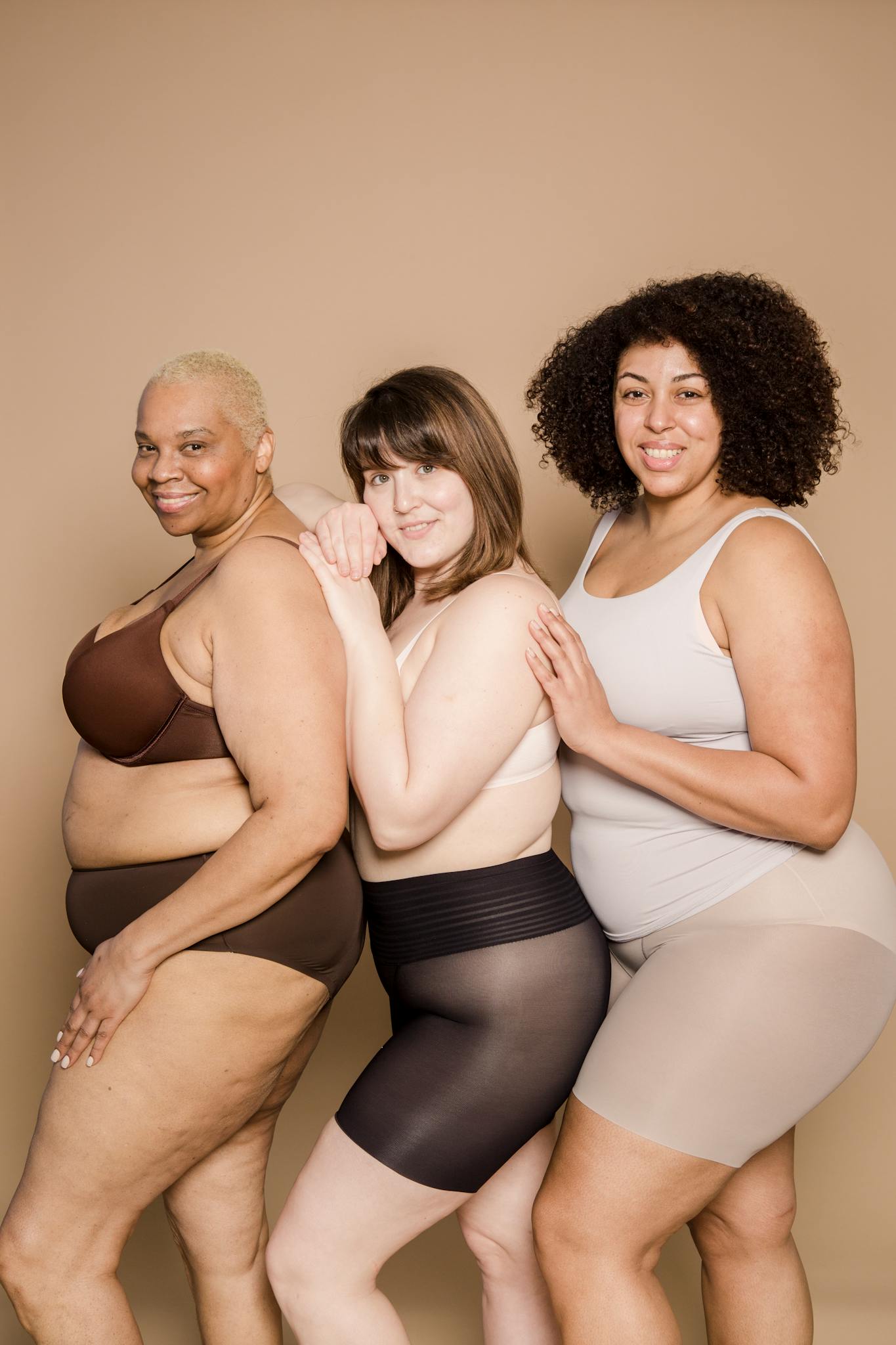 Group of cheerful overweight diverse female models wearing lingerie standing close and looking at camera on beige background in light studio